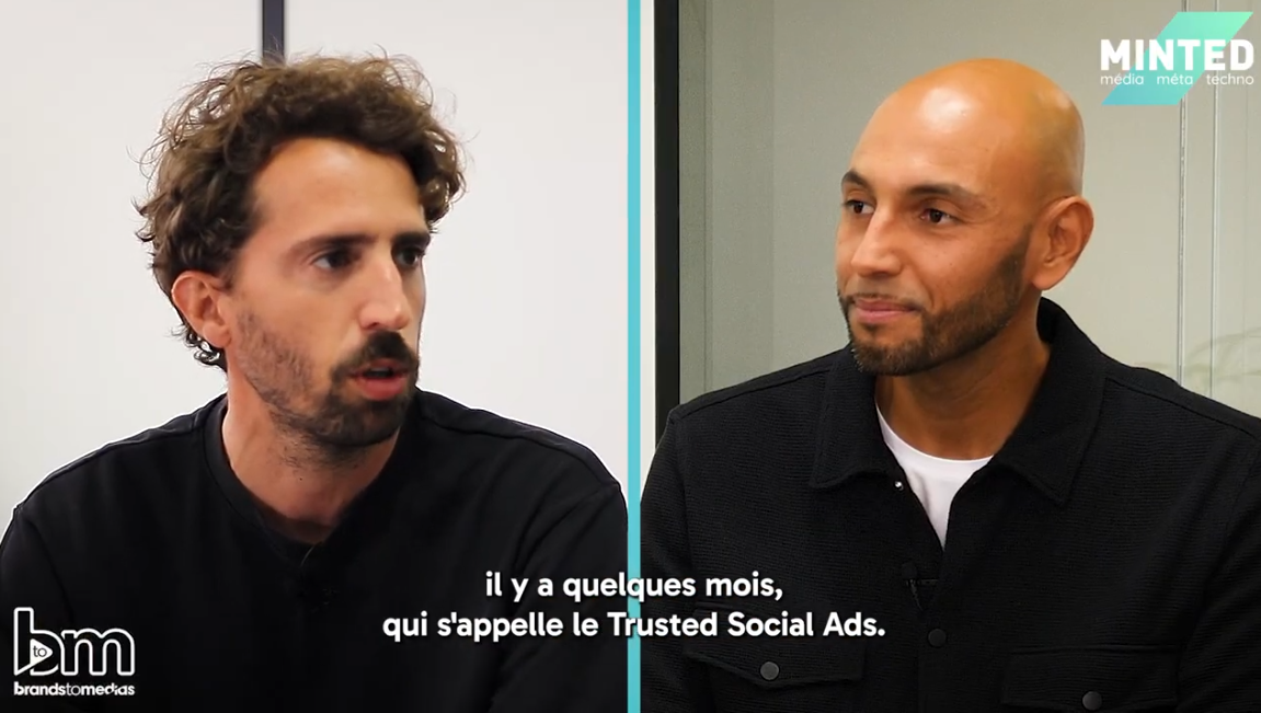 The image is a screenshot from an interview, split into two sections by a vertical light blue line. On the left, a man with curly brown hair, a beard, and wearing a black sweater is speaking with a serious expression. On the right, a bald man with a well-groomed beard, dressed in a black jacket over a white t-shirt, listens attentively with a slight smile. In the top right corner, the logo "MINTED" appears with the words "media," "meta," and "techno." In the bottom left corner, another logo, "bm," followed by "brandstomeckas," is displayed. French subtitles indicate that the conversation is about "Trusted Social Ads." The background is minimalistic, featuring a white wall and a glass partition, giving the setting a professional yet casual tone.